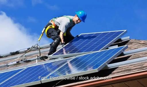 Das Foto zeigt einen Monteur in Arbeitskleidung, gesichert durch einen Gurt und mit blauem Schutzhelm auf dem Kopf, auf einem roten Dach vor blauem Himmel, der ein PV-Modul installiert.