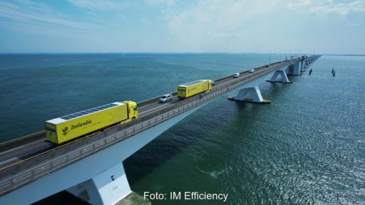 Auf dem Foto ist eine weitläufige Brücke über ein Meer zu sehen, die von links unten schräg nach rechts oben in den Horizont läuft. Auf der Brücke fahren neben PKWs zwei gelbe Trucks mit PV auf dem Dach.