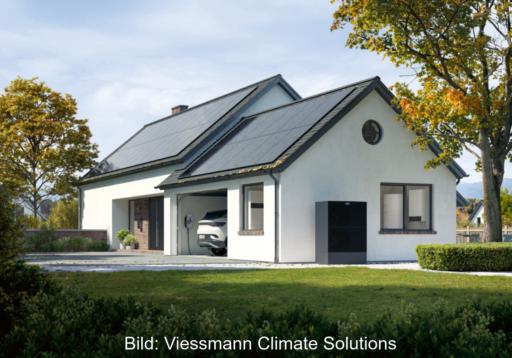 Das Bild zeigt ein weißes Einfamilienhaus, schräg von der Seite, rechts und links Bäume, mit Blick aus dem Grün, in der Garage ein PKW mit Wallbox und vor der Garage eine Wärmepumpe.