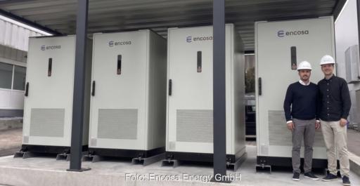 Zwei Personen mit Schutzhelmen neben vier großen Batteriespeichern.