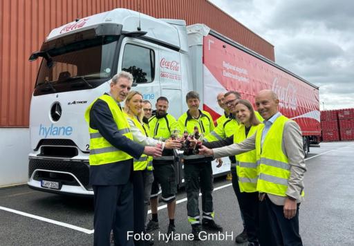 Mehrere Personen in neongelben Warnwesten stehen vor einem batterieelektrischen Mercedes-Benz eActros 600 mit hylane- und Coca-Cola-Schriftzug an der Kabine und stoßen vor dem Fahrzeug mit Getränkeflaschen an.