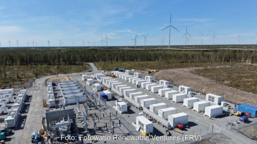 Batteriespeichersysteme auf Beton mit Umspannanlage auf einem Freigelände, im Hintergrund Windkraftanlagen.