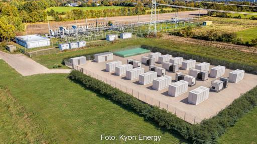 Luftaufnahme eines Batteriespeichers mit mehreren Container-Modulen auf einem eingezäunten Gelände neben einem Umspannwerk. Die Anlage ist von landwirtschaftlichen Flächen umgeben.