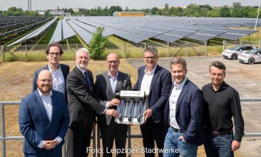 Sieben Personen stehen vor den aufgeständerten CPC-Vakuumröhrenkollektoren einer Solarthermieanlage und halten gemeinsam ein Kollektorelement als symbolisches Übergabeobjekt. Im Hintergrund sind die Kollektorreihen der Anlage sowie ein Industriegebäude zu sehen.