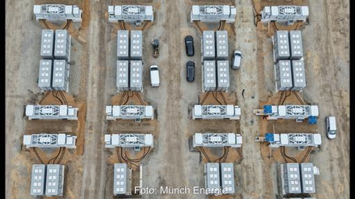 Luftaufnahme eines Großbatteriespeichers auf einer Baustelle. Mehrere Reihen von Batteriecontainern und technischen Umrichtereinheiten sind auf einem geschotterten Gelände angeordnet. Dazwischen stehen mehrere Fahrzeuge.
