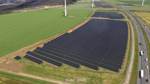 Luftaufnahme eines Solarparks entlang eines Autobahnabschnitts. Mehrere große Photovoltaikfelder liegen neben der Fahrbahn, im Hintergrund sind Windenergieanlagen und landwirtschaftliche Flächen zu sehen.