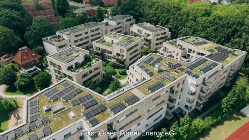 Luftaufnahme einer mehrteiligen Wohnanlage mit begrünten Flachdächern und zahlreichen Photovoltaikmodulen. Die Solaranlagen sind auf mehreren Gebäuden installiert und von grünen Bäumen umgeben.