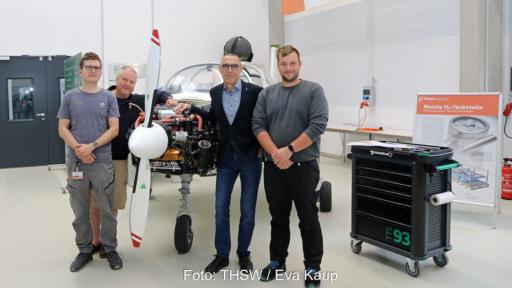 Vier Mitglieder des THWS-Projektteams stehen in einer Werkhalle vor dem zweisitzigen Reisemotorsegler „Taifun 17 H₂“. Die Verkleidung des Flugzeugmotors ist geöffnet und gibt den Wasserstoff-Elektroantrieb frei. Rechts im Bild stehen ein Werkstattwagen und ein Roll-up zur mobilen Wasserstoff-Tankstelle.