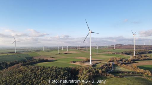 Luftaufnahme eines Windparks in einer landwirtschaftlich geprägten Hügellandschaft. Mehrere Windenergieanlagen stehen zwischen Feldern und Waldflächen. Im Vordergrund ist eine moderne Windkraftanlage zu sehen, im Hintergrund weitere Anlagen.