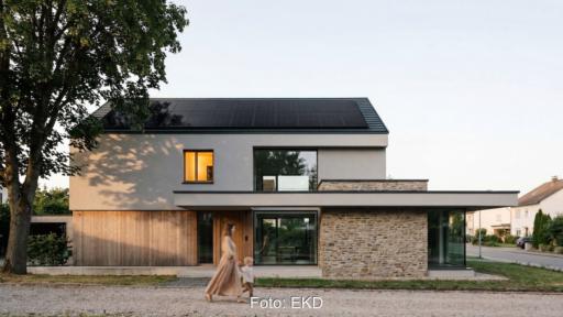Modernes zweigeschossiges Einfamilienhaus mit einer Photovoltaikanlage auf dem geneigten Dach. Vor dem Gebäude läuft eine erwachsene Person mit einem Kleinkind vorbei. Links steht ein großer Baum, im Hintergrund ist eine Wohnstraße zu sehen.