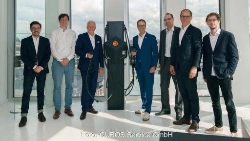 Sieben Männer stehen in einem hellen Büro neben einer schwarzen Ladesäule für Elektrofahrzeuge und blicken in die Kamera. Zwei Personen halten Ladekabel in der Hand. Im Hintergrund sind bodentiefe Fenster mit Blick auf eine Stadt zu sehen.