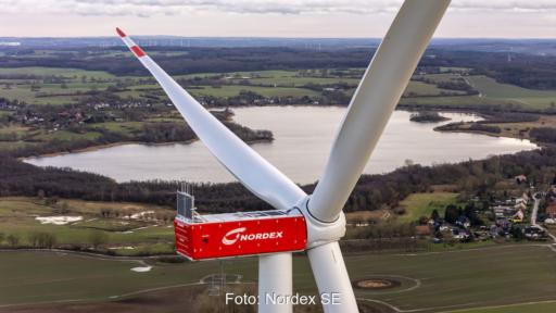 Luft-Nahaufnahme einer Windenergieanlage der Nordex Group mit roter Maschinenhausverkleidung und drei Rotorblättern. Im Hintergrund sind eine Seenlandschaft, Felder, Ortschaften und weitere Windenergieanlagen zu sehen.