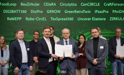 Mehrere Projektbeteiligte stehen auf einer Bühne und präsentieren den Förderbescheid für das Projekt DIGIBATT360. Im Hintergrund sind die Namen weiterer GreenTech-Förderprojekte an einer grünen Wand eingeblendet.