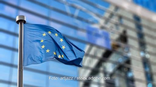 Eine EU-Flagge weht vor einem modernen Glasgebäude in Brüssel, das im Hintergrund unscharf zu sehen ist.