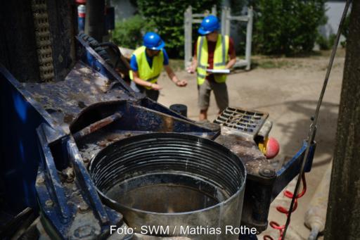 Nahaufnahme einer Brunnenbohrung auf einer Baustelle. Im Vordergrund ist das Bohrgerät mit einem Bohrrohr zu sehen, im Hintergrund stehen zwei Personen mit Schutzhelmen und Warnwesten auf dem Gelände des Pilotprojekts.