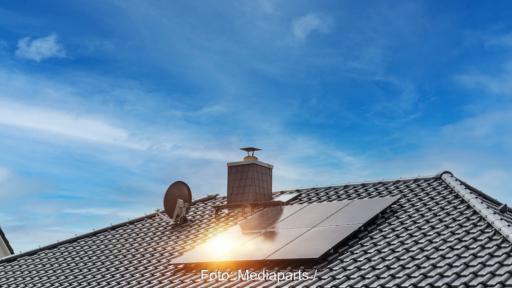 Mehrere schwarze Solarmodule sind auf dem geneigten Ziegeldach eines Einfamilienhauses installiert. Im Hintergrund sind ein Schornstein, eine Satellitenschüssel und ein blauer Himmel mit Wolken zu sehen. Sonnenlicht spiegelt sich auf einem der Module.