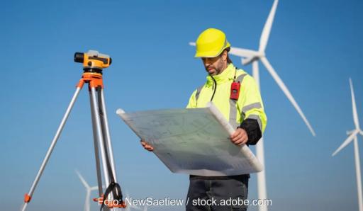 Ein Mann mit gelber Warnweste und gelbem Schutzhelm neben einem Vermessungsgerät hält einen Plan in den Händen. Im Hintergrund stehen mehrere Windenergieanlagen vor einem blauen Himmel.