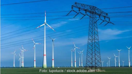 Mehrere Windenergieanlagen hinter Strommast, vor einem blauem Himmel mit leichten Schlieren.