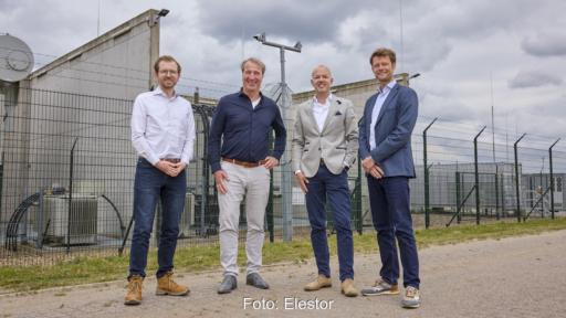 Vier Männer stehen vor einer eingezäunten Hochspannungsanlage mit Transformatoren und weiteren elektrischen Anlagen.