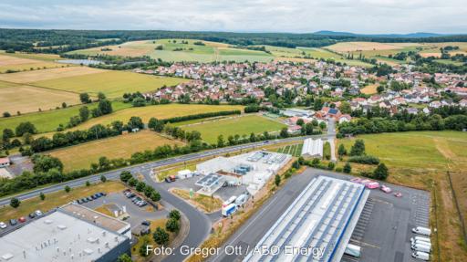 Luftaufnahme des H2-Hubs Hünfeld am Rand von Hünfeld-Michelsrombach. Die technische Anlage liegt zwischen einer Straße, Gewerbegebäuden und Feldern; im Hintergrund sind Wohnhäuser und eine bewaldete Hügellandschaft zu sehen.