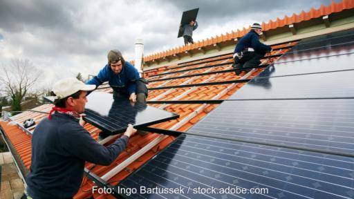 Mehrere Monteure installieren schwarze Photovoltaik-Module auf einem mit roten Ziegeln gedeckten Schrägdach.