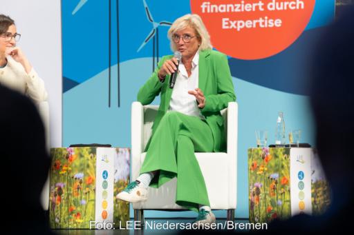 LEE-Vorsitzende Bärbel Heidebroek sitzt im grünen Hosenanzug auf einem Podium und spricht in ein Handmikrofon. Im Hintergrund sind Windenergie-Motive zu sehen.