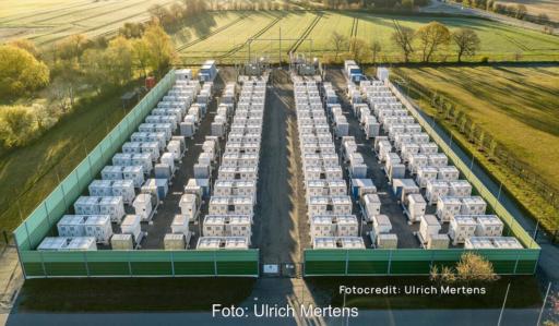 Luftaufnahme des Batteriespeichers Bollingstedt mit zahlreichen weißen Speichercontainern in parallelen Reihen, eine grüne Schutzwand umschließt das Betriebsgelände, umgeben von Feldern und Bäumen.