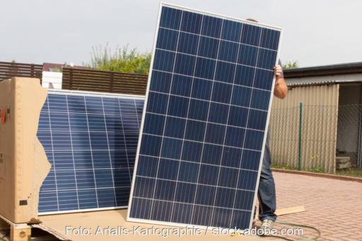 Person hält ein großes PV-Modul neben einem Karton mit weiteren PV-Komponenten.