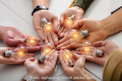 Mehrere Menschen halten kreisförmig ihre Hände nebeneinander auf einer Unterlage und es liegen leuchtende Glühbirnen in den Händen als Zeichen für Energy Sharing.