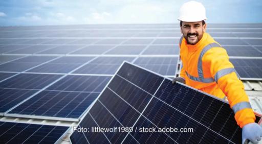 Ein Mann in oranger Arbeitskleidung mit weißem Schutzhelm hält ein Solarmodul vor einem Schrägdach mit weiteren Modulen in den Händen.