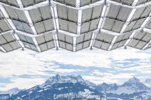 Blick von unten auf die aufgefalteten Photovoltaikmodule eines Solarfaltdachs vor schneebedeckten Bergen.