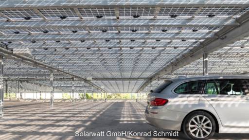 Blick unter eine Parkplatz-Überdachung mit halbtransparenten PV-Modulen, durch die Licht auf den Boden fällt, rechts steht ein Auto.