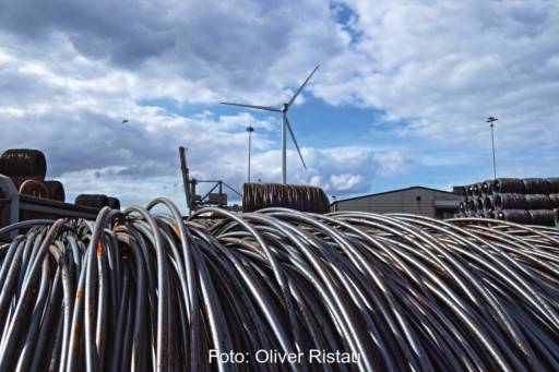 Stahl-Seile im Vordergrund, hinten ein Industriegelände mit einer Windkraftanlage. Erneuerbare Energien für grünen Stahl.