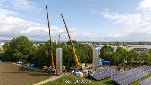 Luftaufnahme zeigt Kräne, die Wärmespeicher bewegen neben einer Freiflächen-PV-Anlage.