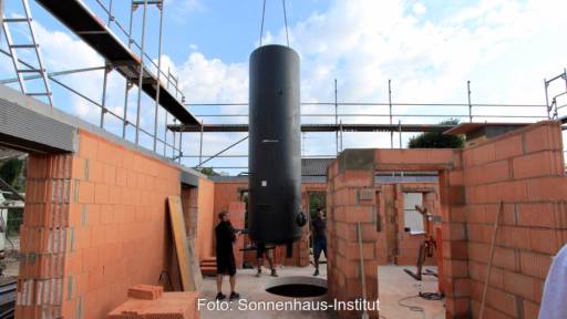 Ein hoher schwarzer Wärmespeicher wird von einem Kran in ein Sonnenhaus im Rohbau hinabgelassen.