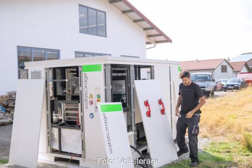 Ein Speicher in einem Gehäuse steht hinter einem Haus, ein Mann steht daneben - der Gewerbe-Speicher in Redox-Flow-Technologie wird aufgebaut.