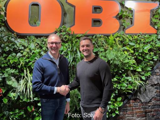 Zwei Männer stehen vor grüner Wand unter Obi-Schriftzug und geben sich die Hand - Partnerschaft von Obi und Soly für PV-Vertrieb