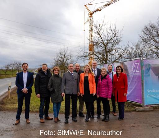 Zehn Personen vor einem Bauzaun, einem Kran an einer Landstraße.