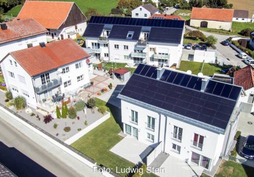 Vier Gebäude im Vordergrund aus der Vogelperspektive. Davon sind zwei mit einem roten Ziegeldach und zwei mit einem dunklen Dach. Diese sind mit Solarthermie-Anlagen bedeckt.