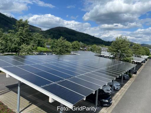 Überdachter Parkplatz mit Solarmodulen vor grünen Bäumen und Hügeln.