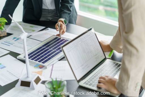 Schreibtisch mit Papieren, Laptop, Solar- und Wind-Modelle und Händen von zwei Personen - Symbol für Planung von Solar-Projekten.