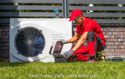 Mann mit rotem Arbeitsanzug und Werkzeugkoffer kniet neben einer Wärmepumpe.