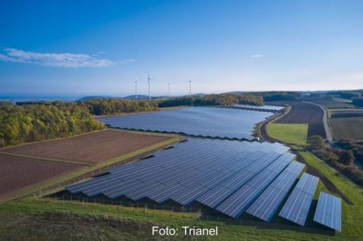 Luftaufnahme eines Freiflächensolarparks neben Äckern unter blauem Himmel.
