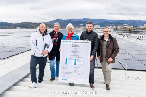 Fünf Personen auf einem Stadiondach mit großer Urkunde vor PV-Anlage.